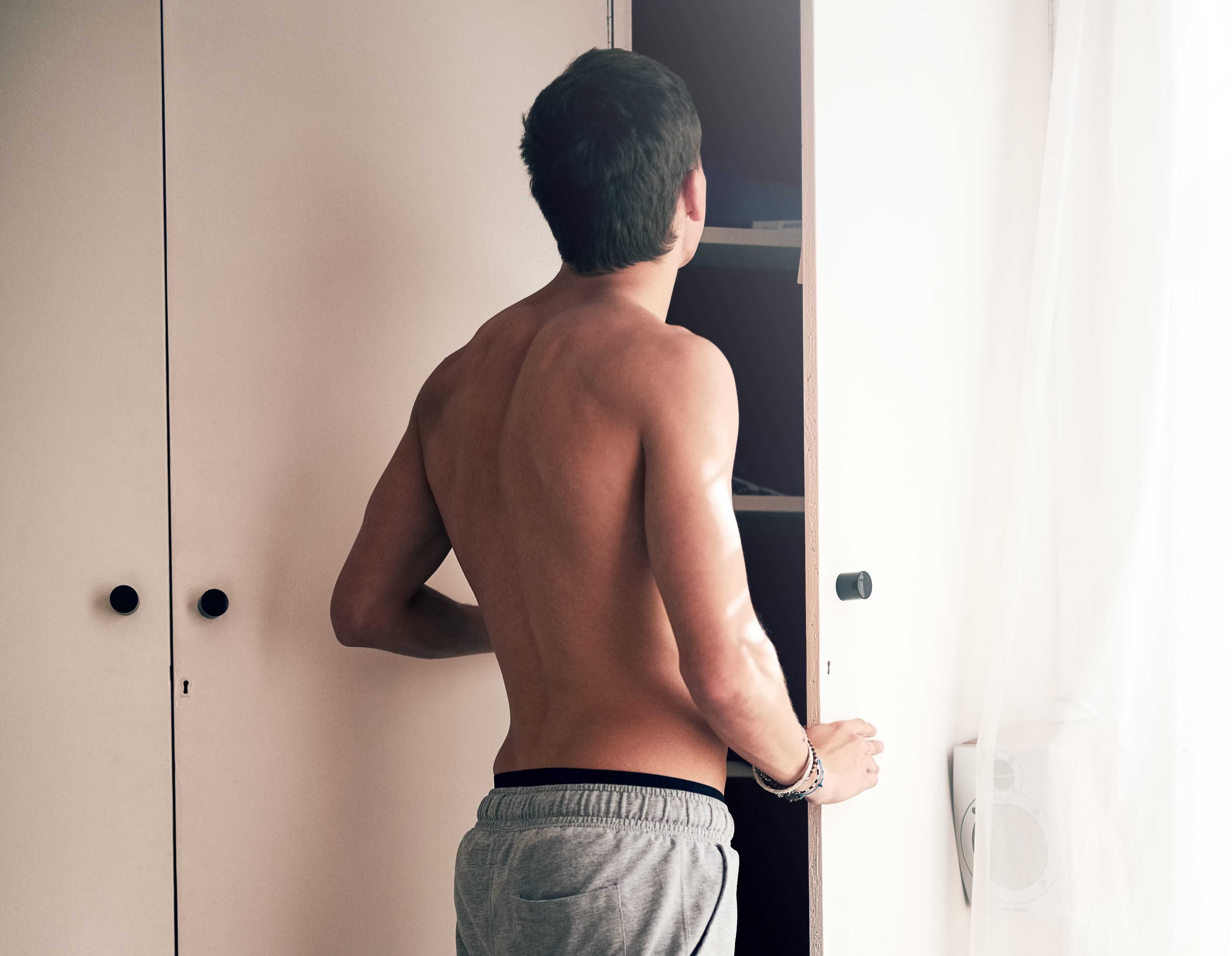 A shirtless person turned away, standing in their closet doorway, and holding the door